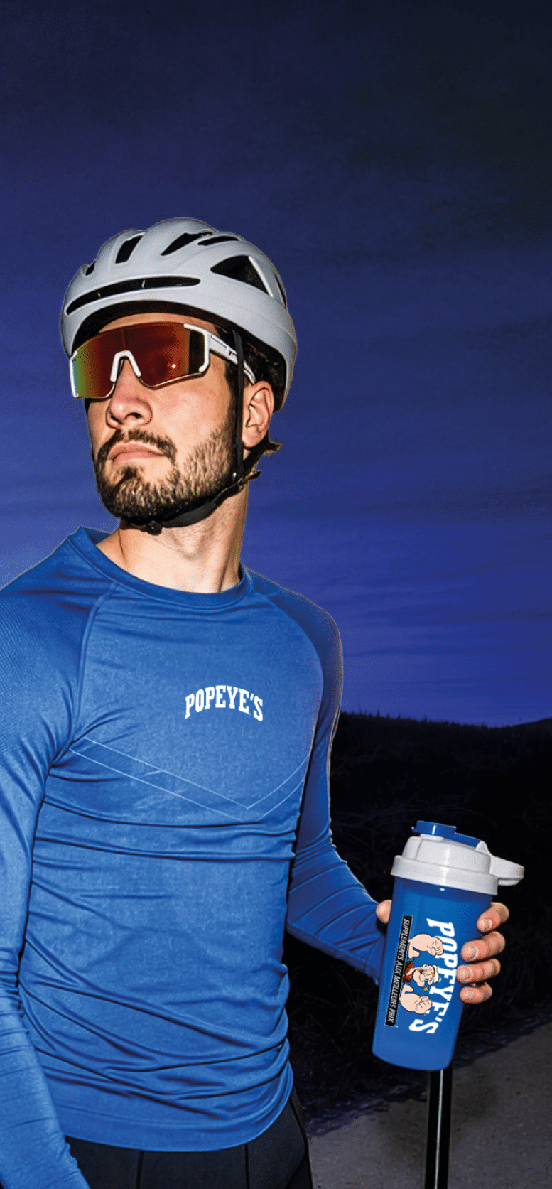 A person wearing a white helmet, sunglasses, and a blue long-sleeve shirt with “POPEYE'S" written on it is holding a blue water bottle with a Popeye cartoon design against a dark outdoor background. Description generated by AI